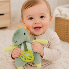 Load image into Gallery viewer, A baby sitting on a white textured rug holding a green plush dinosaur toy with yellow accents in a bright nursery setting.