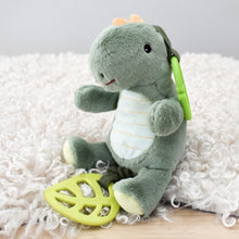 Load image into Gallery viewer, A plush green dinosaur toy sitting on a white shag rug, holding a yellow-green leaf-shaped teether in one foot and wearing a small orange crown.