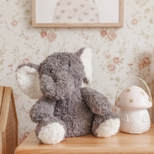 Load image into Gallery viewer, A plush gray elephant toy with white ears, trunk, and feet sits on a wooden table in a nursery setting, next to a white mushroom-shaped night light against a floral wallpaper background.